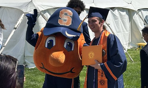 Graduate taking a photo with Otto the Orange outside durning 2023 Commencement.