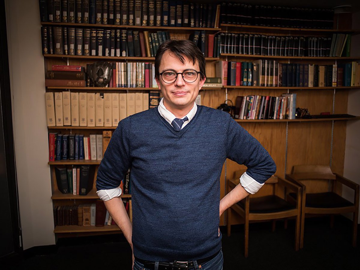 Chris Forster in a blue sweater and tie in front of a wall of a books